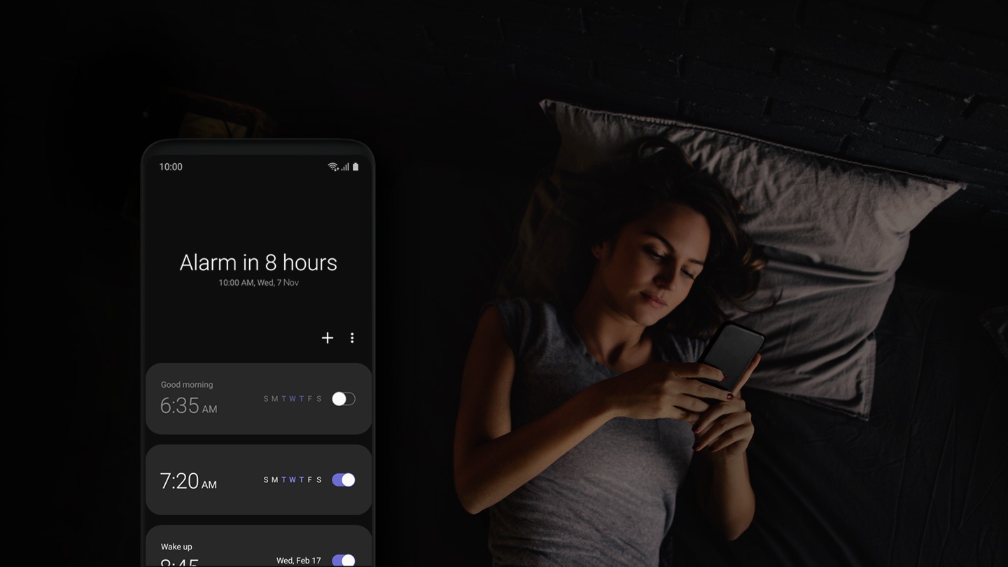 Image of woman lying in bed holding a smartphone with another smartphone in front with One UI Clock application onscreen