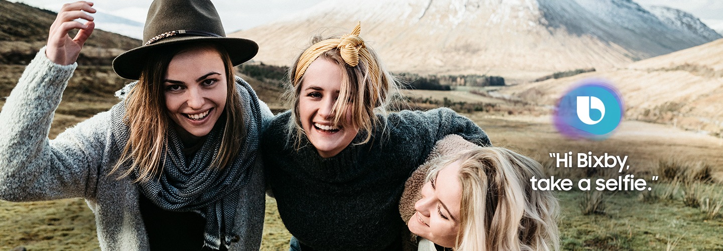 Three women standing on the mountain, putting arms around eath other's shoulders and laughing together. On the right, a bixby icon and text of "Hi Bixby, take a selfie" are shown.