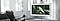 A Samsung lifestyle TV sits on a three-legged stand in front of a black leather armchair in a white room with large windows. There is an image of a large white bird on the screen.