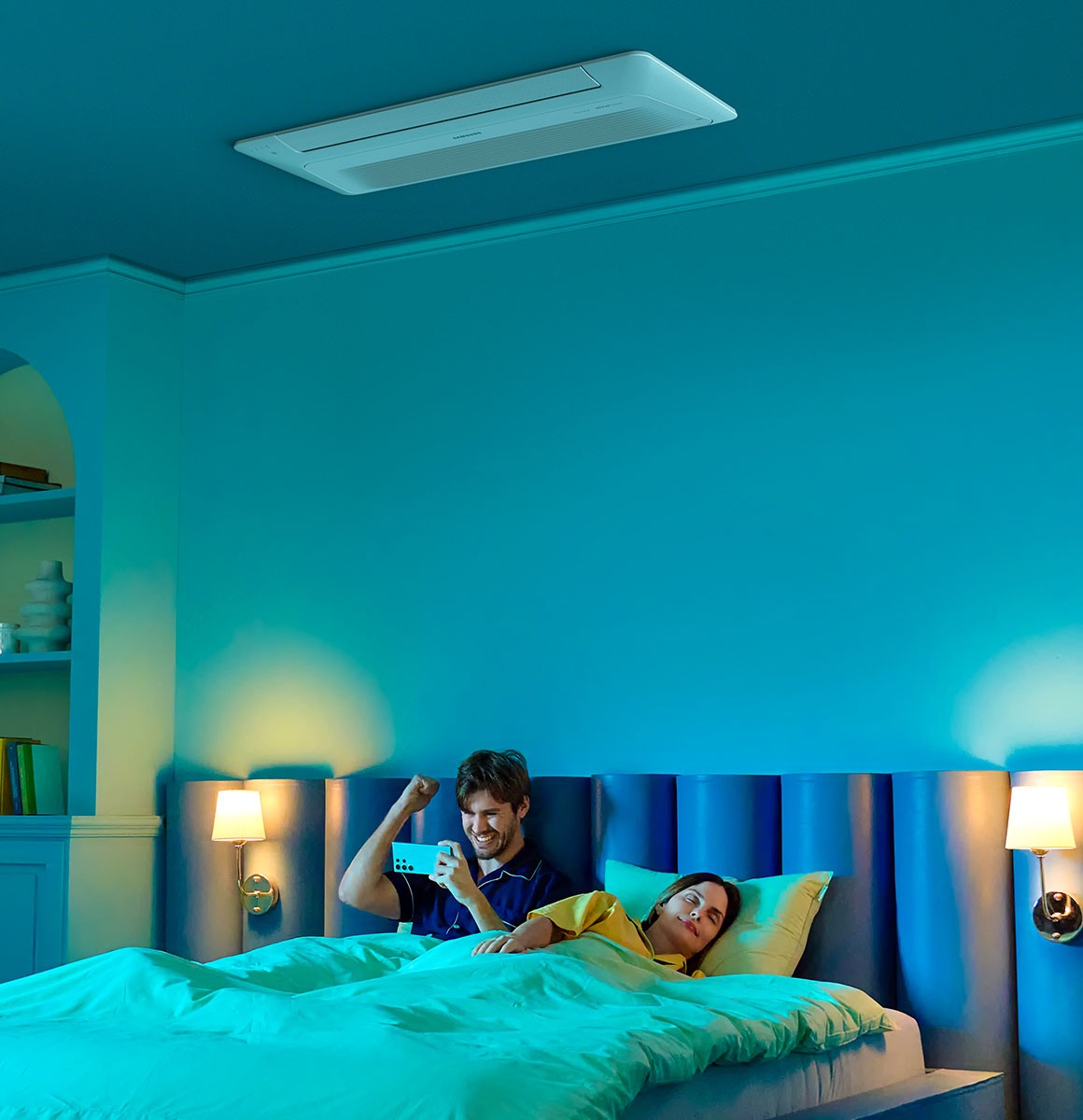 A man enjoys content on his phone in bed while a woman sleeps next to him. Above them is a 1Way Cassette AC.