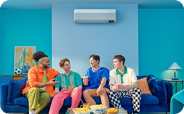Four young men with colorful attire chat and enjoy snacks on a couch. A WindFree AC is mounted on a light blue wall.