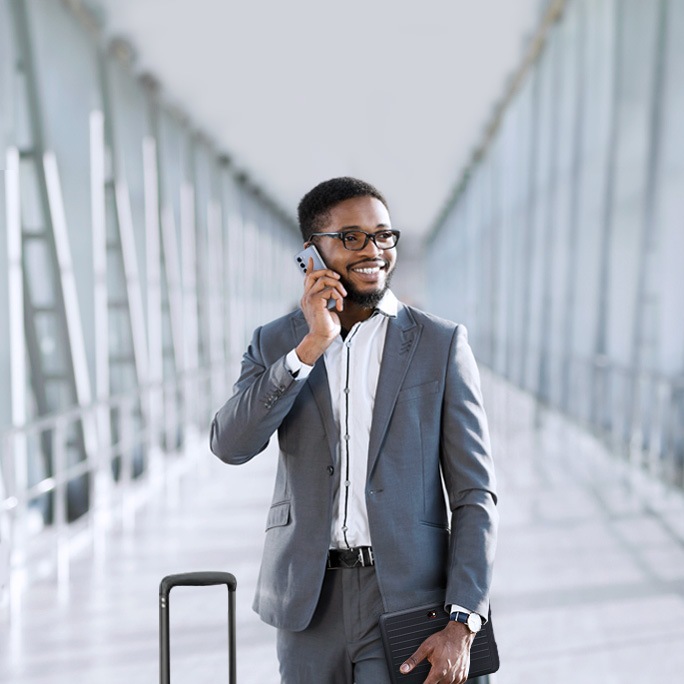 A man in a business suit is walking through an airport terminal, talking on his phone and smiling. He is pulling a suitcase with one hand and holding a tablet with the other. The terminal is spacious and modern, with large windows allowing natural light to flood in. The text on the image reads "Essentials for work and travel." The scene conveys a sense of professionalism and readiness for business travel.