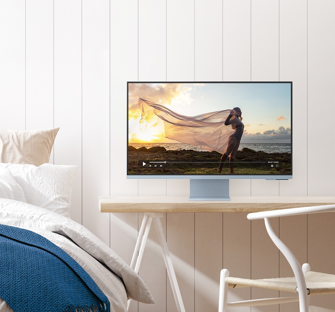 There is a blue monitor on top of a different desk. And a video with a lady standing in the nature is being played on the monitor. A desk chair is shown in front of the desk. To the left of a desk is a bed with pillows and a folded comforter on top.