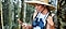 A woman in a wide-brimmed hat with binoculars around her neck checks her phone while leaning against a tree in the woods.