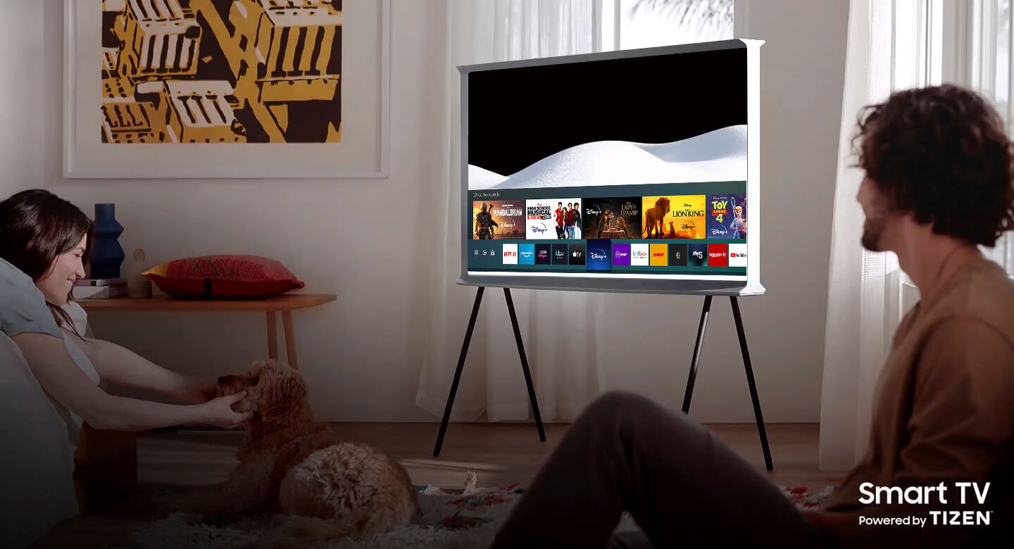 A man and a woman are enjoying their time together in a living room.  A Cloud White The Serif sits in the center of the room with the screen Samsung Smart Hub open.The Serif TV is Smart TV powered by Tizen.