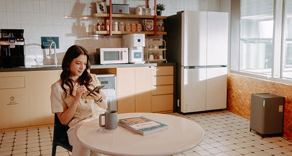 A woman is sitting in a kitchen equipped with various Samsung appliances and taking notes on her cell phone.