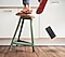 A closeup view shows a person's feet standing precariously on a wobbly kitchen stool. The stool is tilting to the right, with only one of its three legs on the floor, and the person's smartphone is falling to the ground in mid-air.