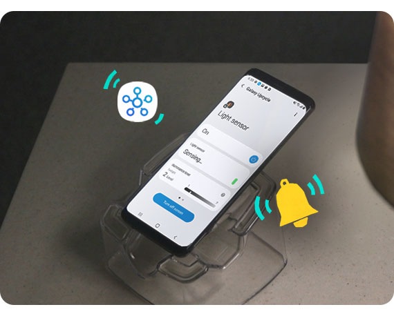 Um telemóvel Samsung Galaxy é mostrado em grande plano, voltado para baixo e ligeiramente inclinado para a direita. O telemóvel Samsung Galaxy está em cima de um suporte de telemóvel transparente. Existe uma ilustração amarela com formato de sino e a ilustração do ícone do SmartThings, todos mostrando as diferentes funções para as quais podem ser utilizados os antigos dispositivos Samsung Galaxy.