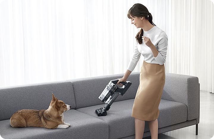 Një grua po pastron divanin me një fshesë jet 75. Në të majtë është një qen.