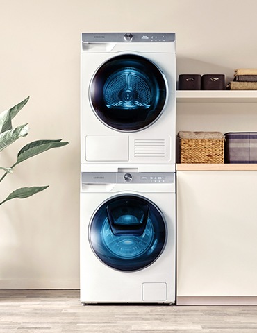 Kombinimi i makinës larëse dhe tharëse njëra mbi tjetrën është i vendosur ndërmjet një bime të gjelbërt dhe dollapit me sirtare.