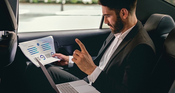 En el asiento trasero de un automóvil, un hombre con traje está revisando documentos comerciales en su mano con un Galaxy Book en su regazo.