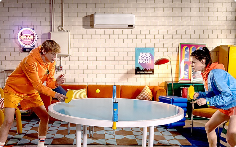 A WindFree AC is mounted on a white tiled wall. A man and a woman in colorful athletic wear are playing Ping-Pong on a round white table at the center of the room.