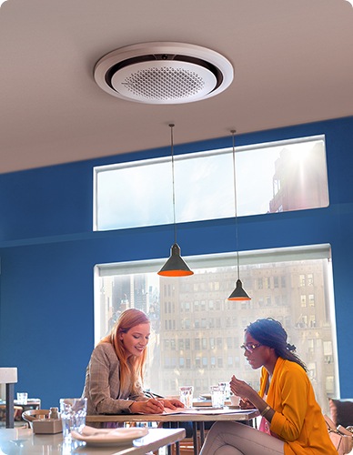 Two women are sitting at a table in front of a large window in a restaurant. One woman is writing on a piece of paper while the other woman watches. A 360 Cassette AC is mounted on the ceiling above.