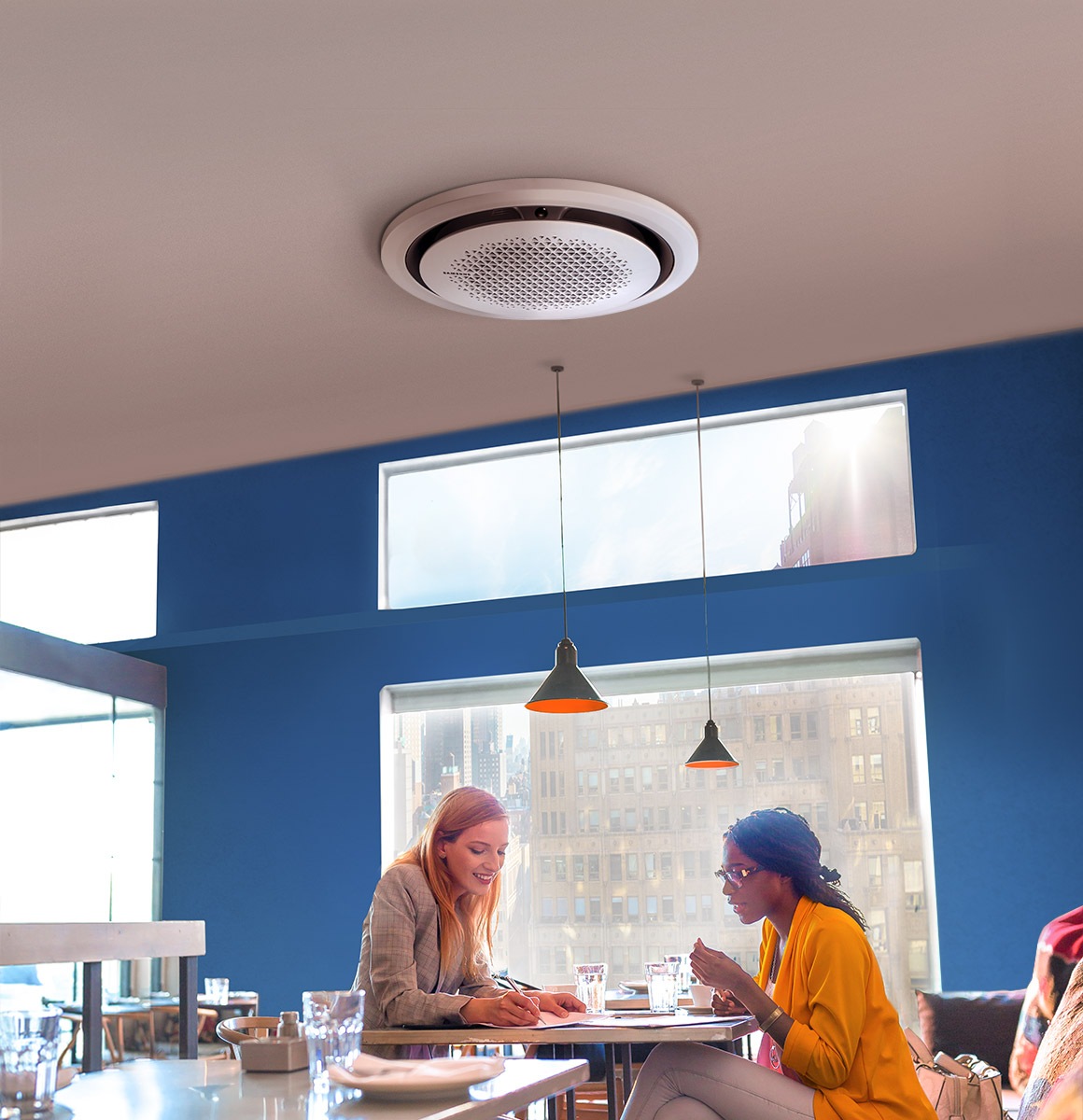 Two women are sitting at a table in front of a large window in a restaurant. One woman is writing on a piece of paper while the other woman watches. A 360 Cassette AC is mounted on the ceiling above.