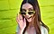 Una mujer sonriendo está de pie frente a una pared verde brillante, asomándose desde sus anteojos de sol de color verde brillante.