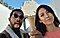 Un hombre y una mujer se toman una selfie juntos en un jardín de esculturas. El hombre está usando anteojos de sol y sacando la lengua mientras la mujer sonríe a la cámara. El hombre también sostiene un cono de helado mientras la roca en el fondo parece estar apoyada sobre él gracias a una perspectiva forzada.