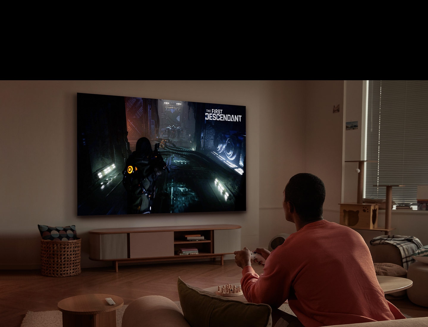 Ein Mann spielt ein Videospiel auf einem großen Fernsehbildschirm im Wohnzimmer mit einem drahtlosen Controller. Das Logo des Spiels 'First Descendant' befindet sich oben auf dem Bildschirm.