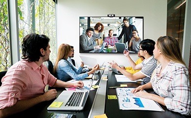 Many people are sitting in the conference room, and they are having a video conference while watching the QxB installed in the front.