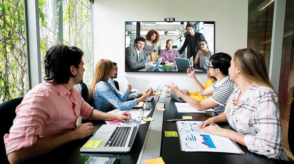 Many people are sitting in the conference room, and they are having a video conference while watching the QxB installed in the front.