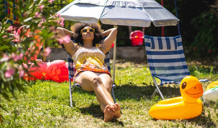 Žena sa sunčanim naočalama opušta se u svom dvorištu, okružena suncobranom za plažu, patkom na napuhavanje i baštenskom stolicom.