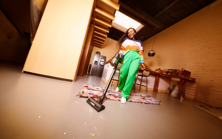 Een vrouw in een keuken die de vloer stofzuigt met een Samsung Jet Bot. Ze draagt en groene broek en kleurrijke trui.