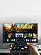 We zien een smartphone in iemands rechterkant met een afstandsbediening gericht op een aan de muur bevestigde Smart TV.