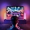 Een persoon die in een kamer met sfeerverlichting zit en een levendig racevideospel speelt op een groot tv-scherm. De kamer is uitgerust met kleurrijke sfeerverlichting voor een betere gamingervaring.
