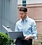 A man in business attire sits outdoors looking at files in his right hand while A Galaxy Book sits on his lap.