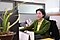 Une femme en tailleur vert est assise à son bureau et regarde l’écran de l’ordinateur. Elle a les cheveux courts et porte des lunettes. Son bureau, sur lequel repose une grande plante, est situé dans un open space.