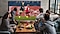 Un groupe de cinq hommes assis autour d'une table basse regardent et applaudissent avec enthousiasme un match de football sur une Super Big TV.