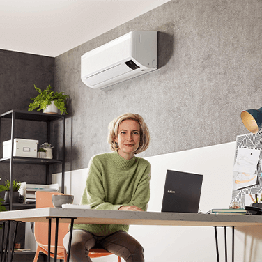 Жанчына сядзіць з ноўтбукам у пакоі з насценным кандыцыянерам.