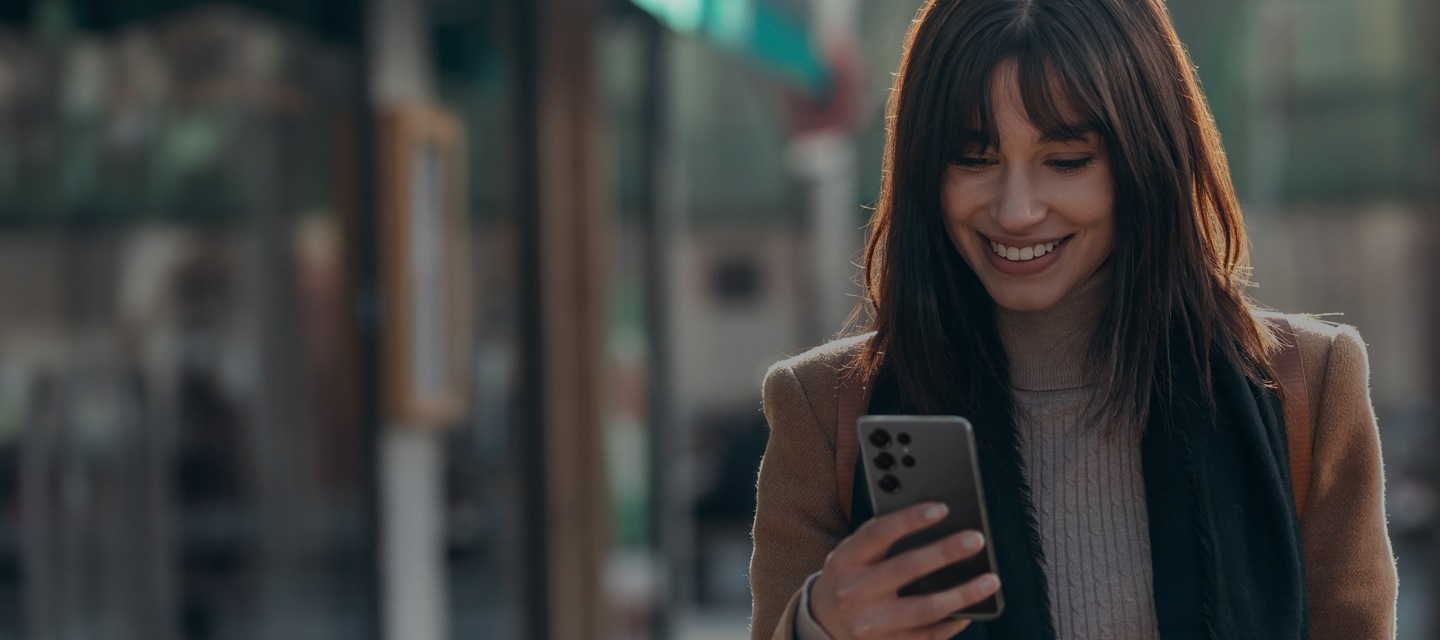 Uma mulher e um homem, cada um ao ar livre em diferentes locais, estão olhando para seus smartphones Galaxy com um sorriso.