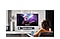 Uma mulher sentada em um sofá em uma sala de estar iluminada assistindo a um vídeo de um show em uma Smart TV no suporte de parede. A mulher ativa o assistente de voz dizendo “Abaixar o volume” no controle remoto da Smart TV que ela está segurando.