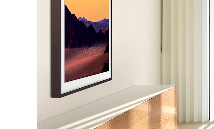 Dentro de uma sala de estar iluminada, a TV The Frame é vista de perto, pela lateral, e pendurada usando a opção de suporte de parede. Há uma imagem de uma cena de montanha ao pôr do sol na tela.