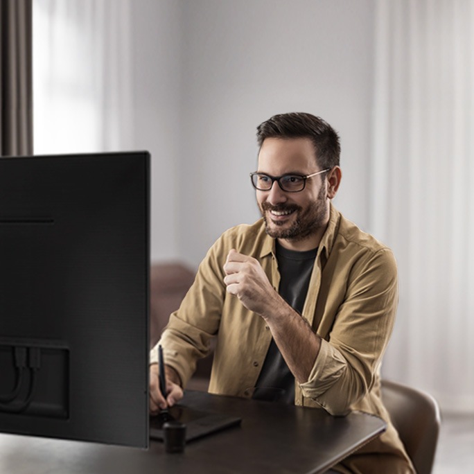 A man with glasses is working at a desk in a modern home office. He is smiling and looking at a large monitor while using a stylus and tablet for his work. The room is well-lit with natural light coming through a window. The text on the image reads "High-end gear, high-end performance." The setting conveys a sense of productivity and comfort, highlighting the advanced technology he is using.