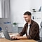 A man in a micro business office working on a laptop and a large monitor.