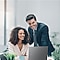 A man and a woman smiling while looking at their laptops in a small business office space.