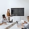 A woman is giving a presentation in front of several people. Her presentation is shared with the people through a large monitor. The mid-size business workers focus on the meeting.