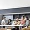 Two groups of business professionals are chatting in a company lounge. Three people are talking while looking at Galaxy smartphones. Two people are looking at Samsung tablets together in the back.