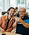 An older and younger man sitting down in a cafe, looking at a Phantom Black Samsung Galaxy S21 phone, smiling.