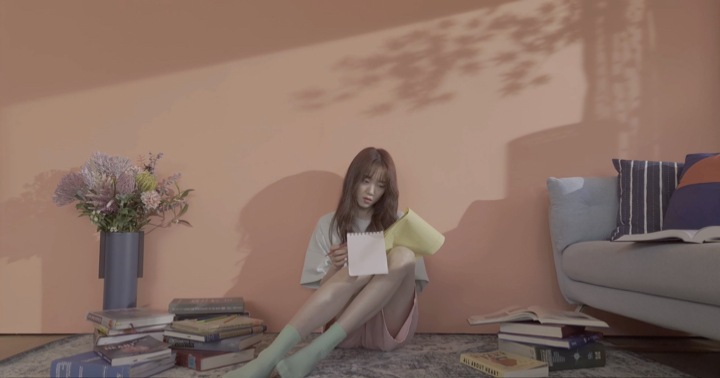 A woman sits on the floor against a wall, surrounded by books and writing in a notepad.