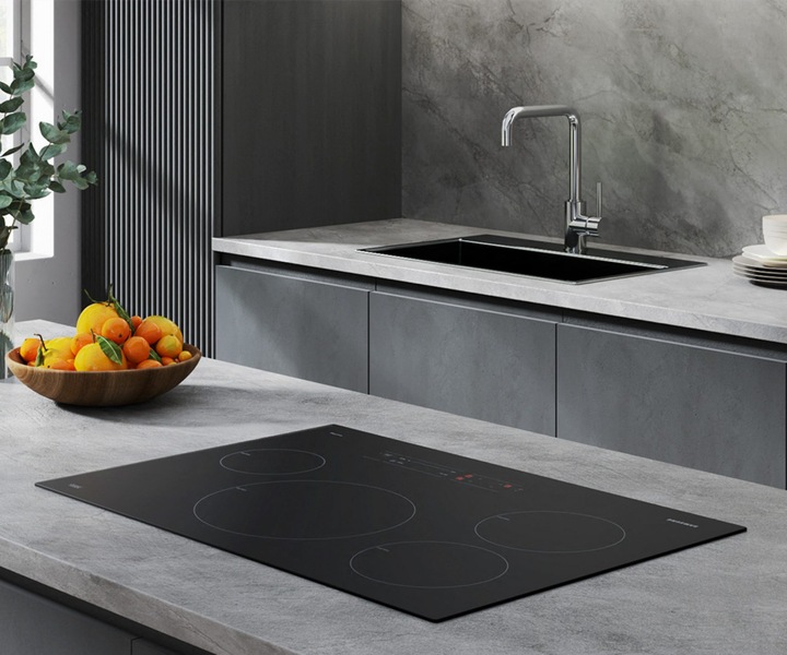 Cuisine moderne avec une plaque de cuisson électrique Samsung élégante sur un comptoir gris, comprenant un bol d’oranges, un bouquet d’eucalyptus dans un vase en verre et un évier avec un robinet au design raffiné.