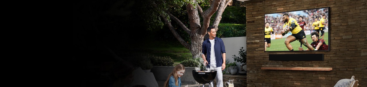 New 2020 Samsung The Terrace soundbar and The Terrace TV are seen on a wall at an angle. And people are preparing for dinner in the backyard .