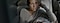 A woman in a beige sweater, sitting on her couch while holding her Galaxy phone. Her body is turned toward the wall and she is looking out the window as her left hand supports her head.