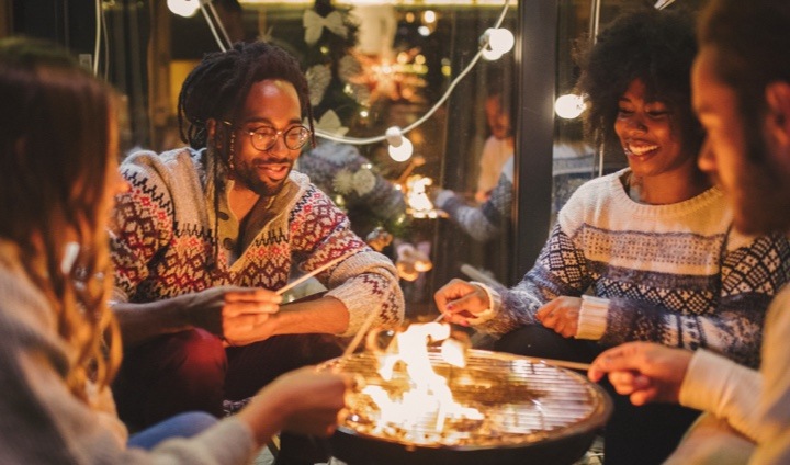 Un groupe de quatre adultes assis à l’extérieur font griller des guimauves autour d’une table à feu.