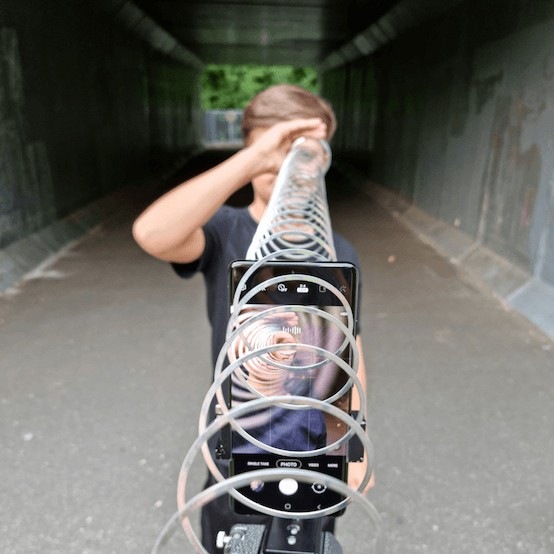 Un Galaxy S21 Ultra 5G posé sur un trépied pointé vers un garçon tenant l’extrémité d’un slinky devant son œil. Le reste du slinky est tendu au-dessus du téléphone vers le trépied et le photographe, créant un effet de spirale sur l’image dans le viseur du téléphone.