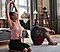 Un homme et une femme sont assis sur le plancher d’un appartement dans des vêtements d’entraînement. La femme s’étire au-dessus de la tête dans une posture de yoga pendant que l’homme regarde.