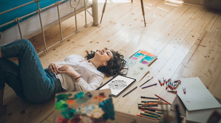 Une femme est allongée sur le dos sur un plancher en bois, avec des marqueurs, des crayons, de la peinture et d’autres fournitures artistiques autour d’elle.