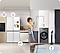 A woman is sitting on the counter of a kitchen that turns into a bathroom counter, merging the two living spaces. She is holding her phone, to which all the appliances are connected, as indicated by white lines and device icons.