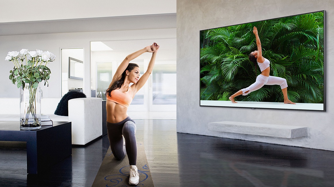 In einem weiß eingerichteten Wohnzimmer macht eine sportlich gekleidete Frau Yoga-Übungen, während ein Yoga-Trainings-Programm auf dem Super Big TV läuft.